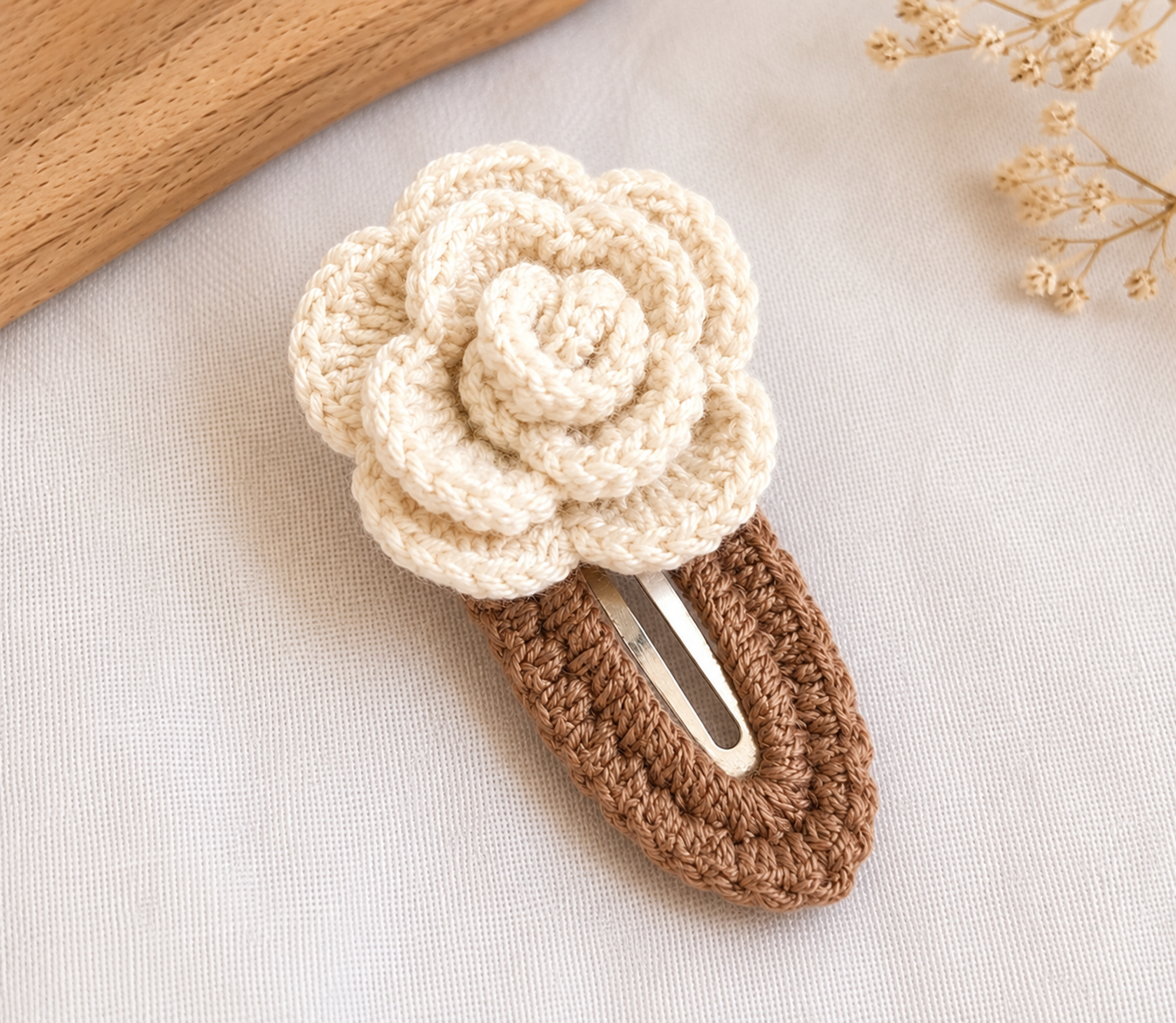 A crocheted hair clip featuring a large cream-colored flower on top of a brown textured base, with a silver metal spring. The clip is styled on a light beige fabric background with a wooden slice and dried flowers as props.