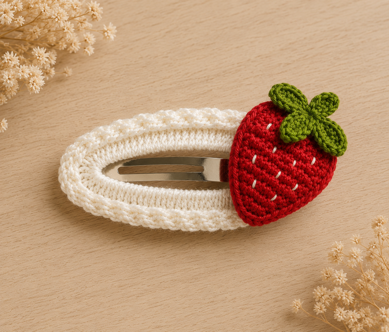 A white crocheted hair clip decorated with a red crocheted strawberry featuring a green leafy top, displayed on a light wood surface with dried flowers in the corners.