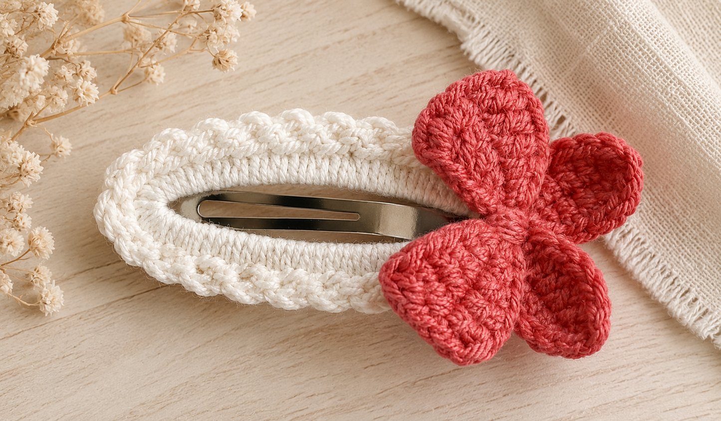 A white crocheted hair clip with a red crocheted bow accent, displayed on a light wood surface with dried flowers and a beige fabric in the background.