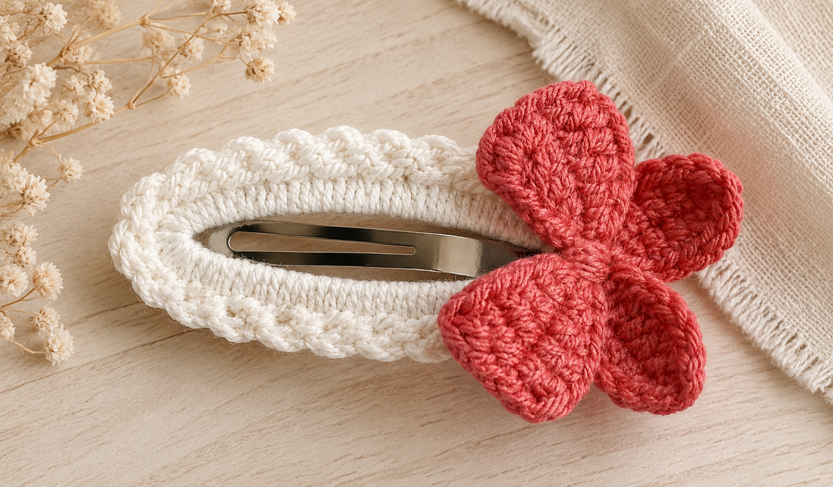A white crocheted hair clip with a red crocheted bow accent, displayed on a light wood surface with dried flowers and a beige fabric in the background.