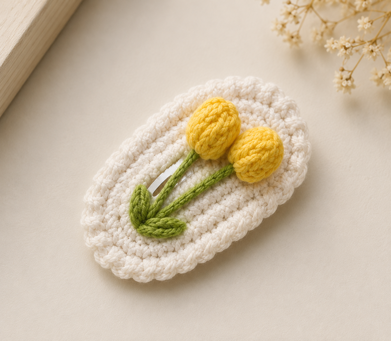 A crocheted oval hair clip in off-white with two yellow tulip flowers and green stems, displayed on a light beige surface with dried flowers and a book corner in the background.