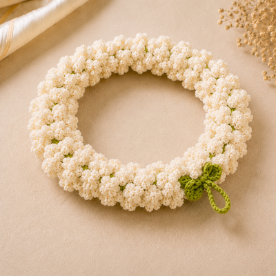 A crocheted floral wreath made of small white flowers with a small green leaf and stem detail, displayed on a beige fabric background.