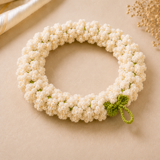 A crocheted floral wreath made of small white flowers with a small green leaf and stem detail, displayed on a beige fabric background.