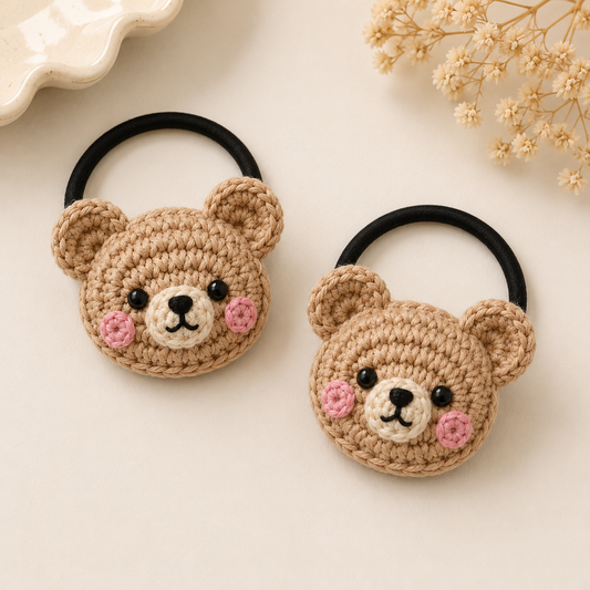 Two crocheted bear-shaped hair ties with black elastic bands, displayed on a light beige background with dried flowers and a ceramic dish edge.