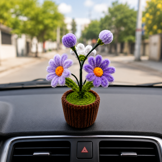 A small handmade flower arrangement in a brown pot sits on a car dashboard with a blurred street scene in the background.