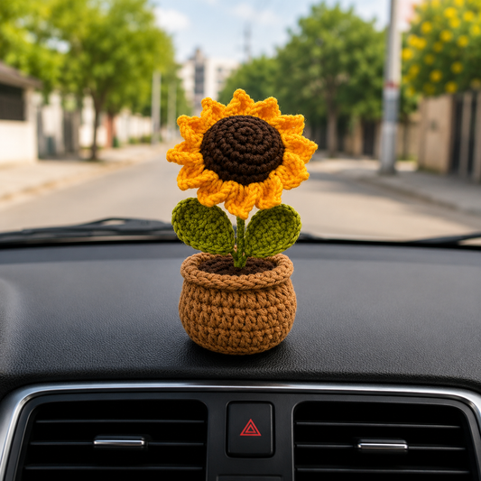 A crocheted sunflower car air freshener sits on a black car dashboard in front of the windshield, with a blurred street scene in the background.