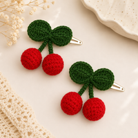 Two cherry-shaped hair clips with red crochet pom-pom cherries and green crochet leaf stems, displayed on a light cream background with decorative lace and dried flowers.