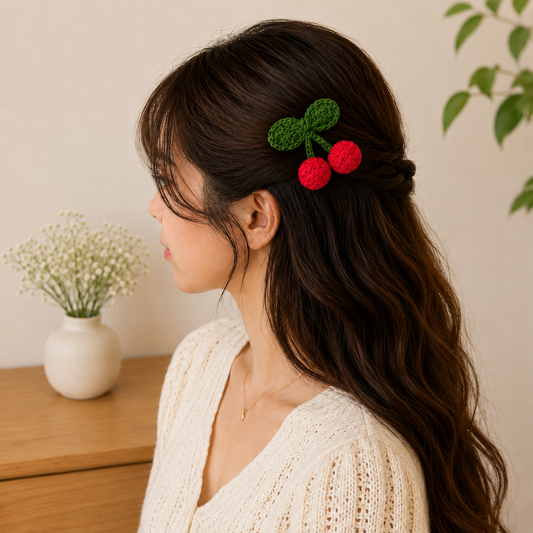 A woman in profile wearing a crocheted hair accessory shaped like two red cherries with green leaves, styled in her dark brown hair. She is wearing a cream-colored knit sweater, with a wooden table, a small white vase of baby's breath, and a green plant in the background.
