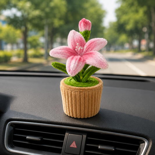 A small pink lily flower ornament in a ribbed tan pot sits on a car dashboard with a blurred road and trees in the background.