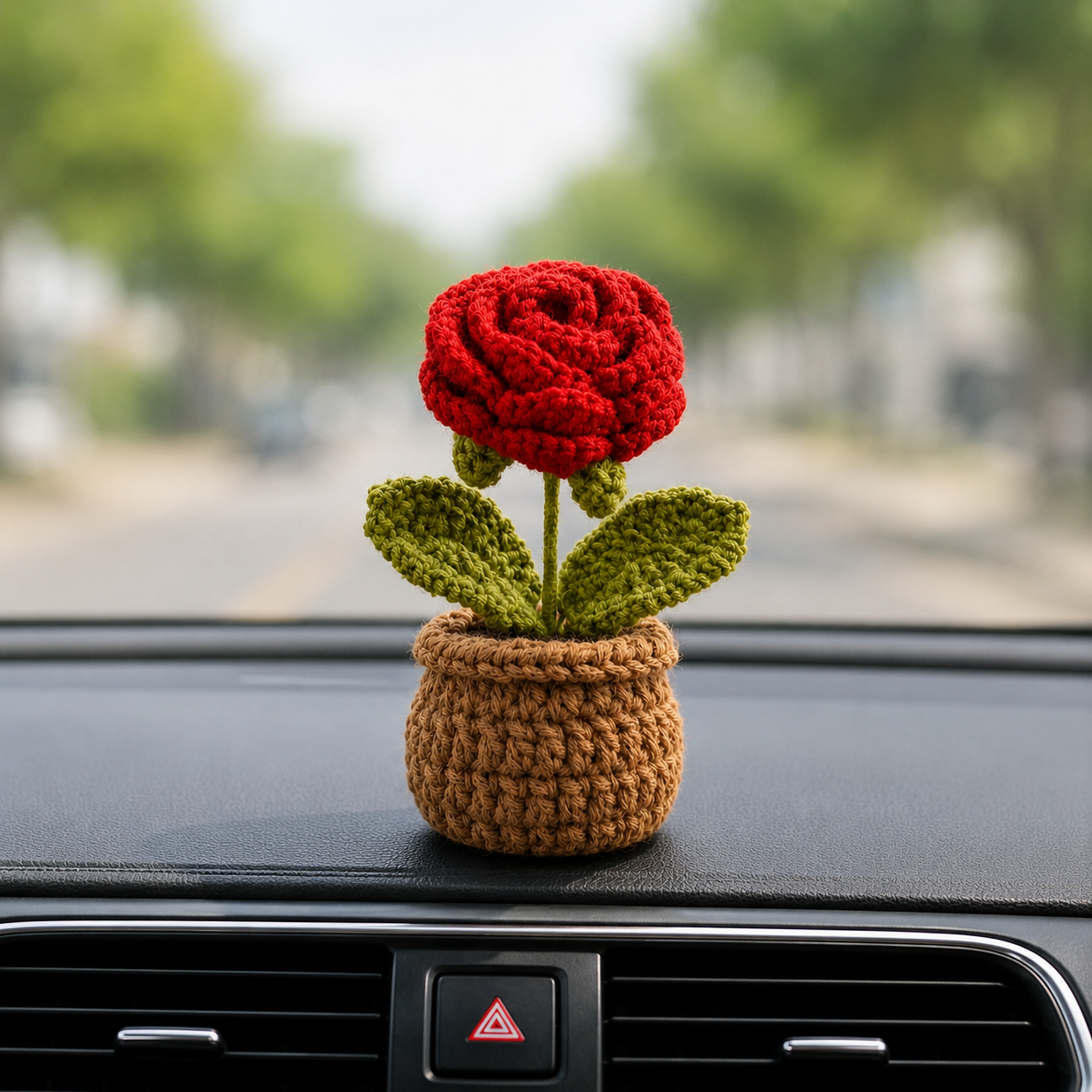A crocheted red rose flower in a small brown pot, placed on a car dashboard with a blurred street background visible through the windshield.