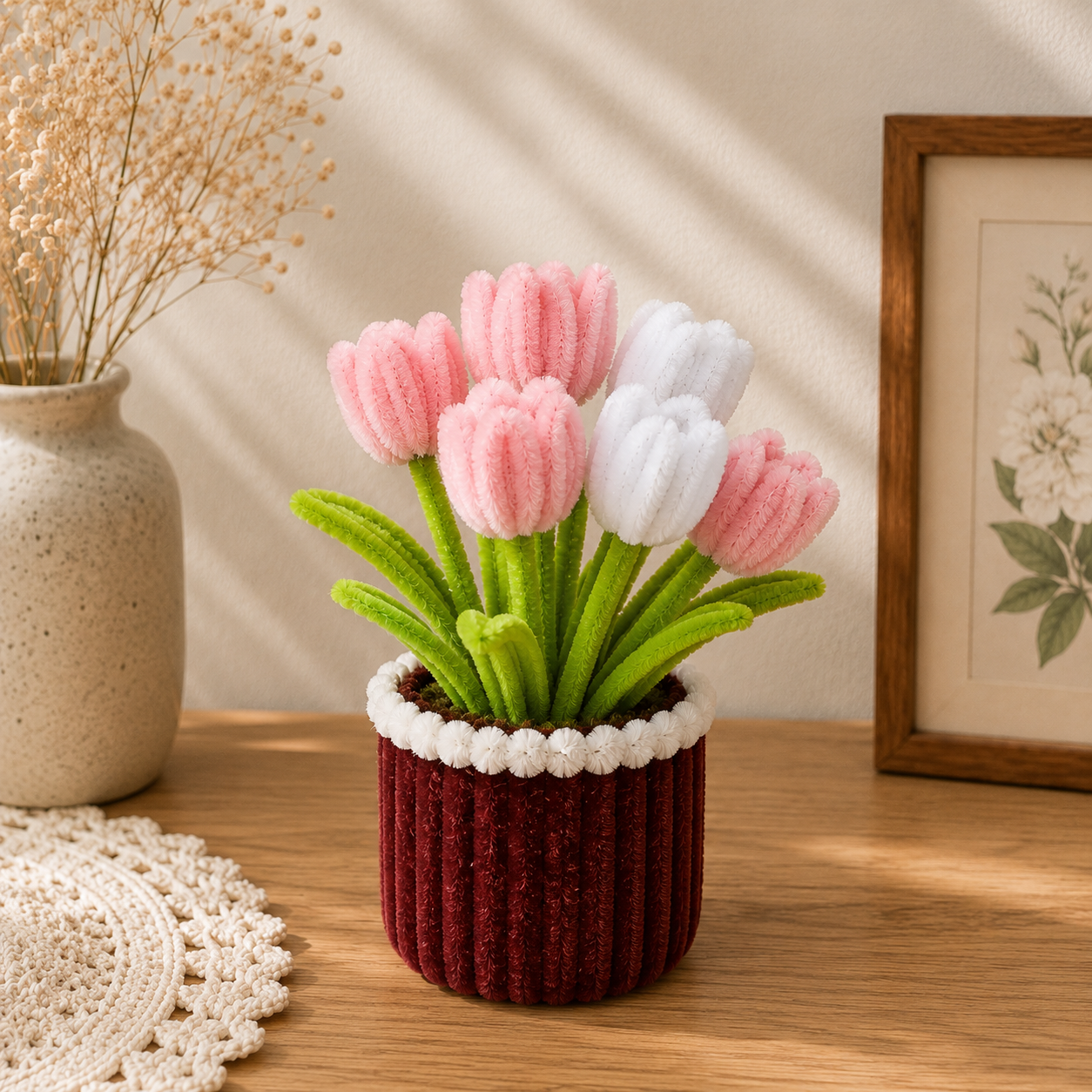 A handmade felt tulip arrangement in a ribbed burgundy pot with a white flower-trim, displayed on a wooden table in a styled home setting with a ceramic vase and framed art in the background.