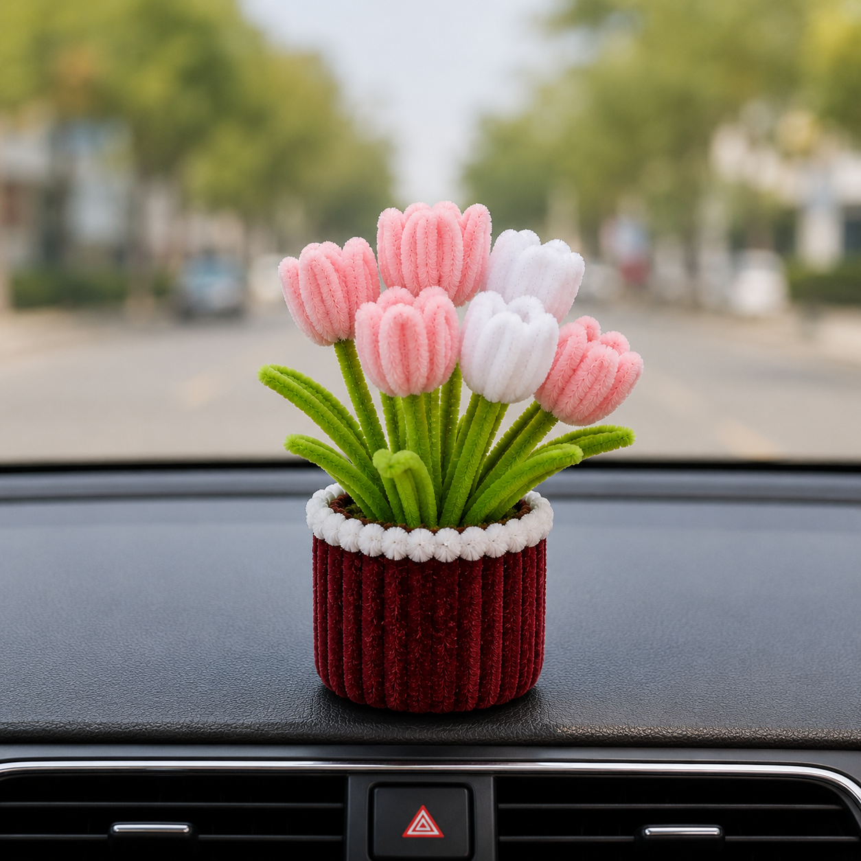 A small decorative flower arrangement in a ribbed red pot with white trim, placed on a car dashboard with a blurred street scene behind it.