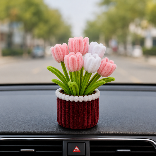 A small decorative flower arrangement in a ribbed red pot with white trim, placed on a car dashboard with a blurred street scene behind it.