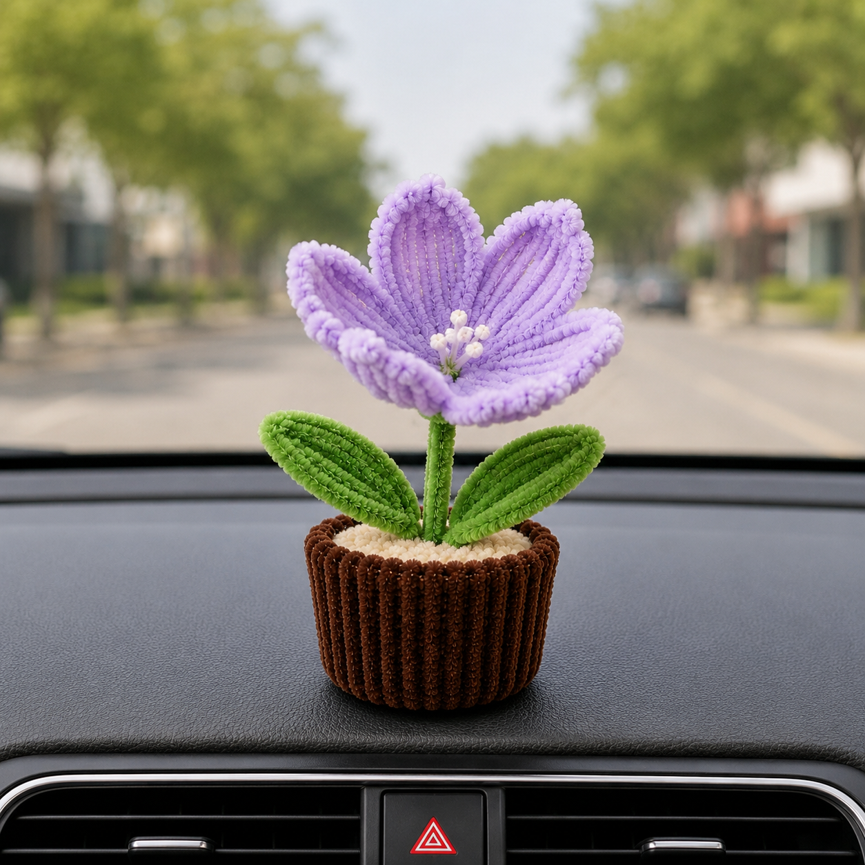 A small crocheted or knitted flower car accessory with a purple bloom, green leaves, and a brown ribbed pot, placed on a car dashboard with a blurred street scene behind it.