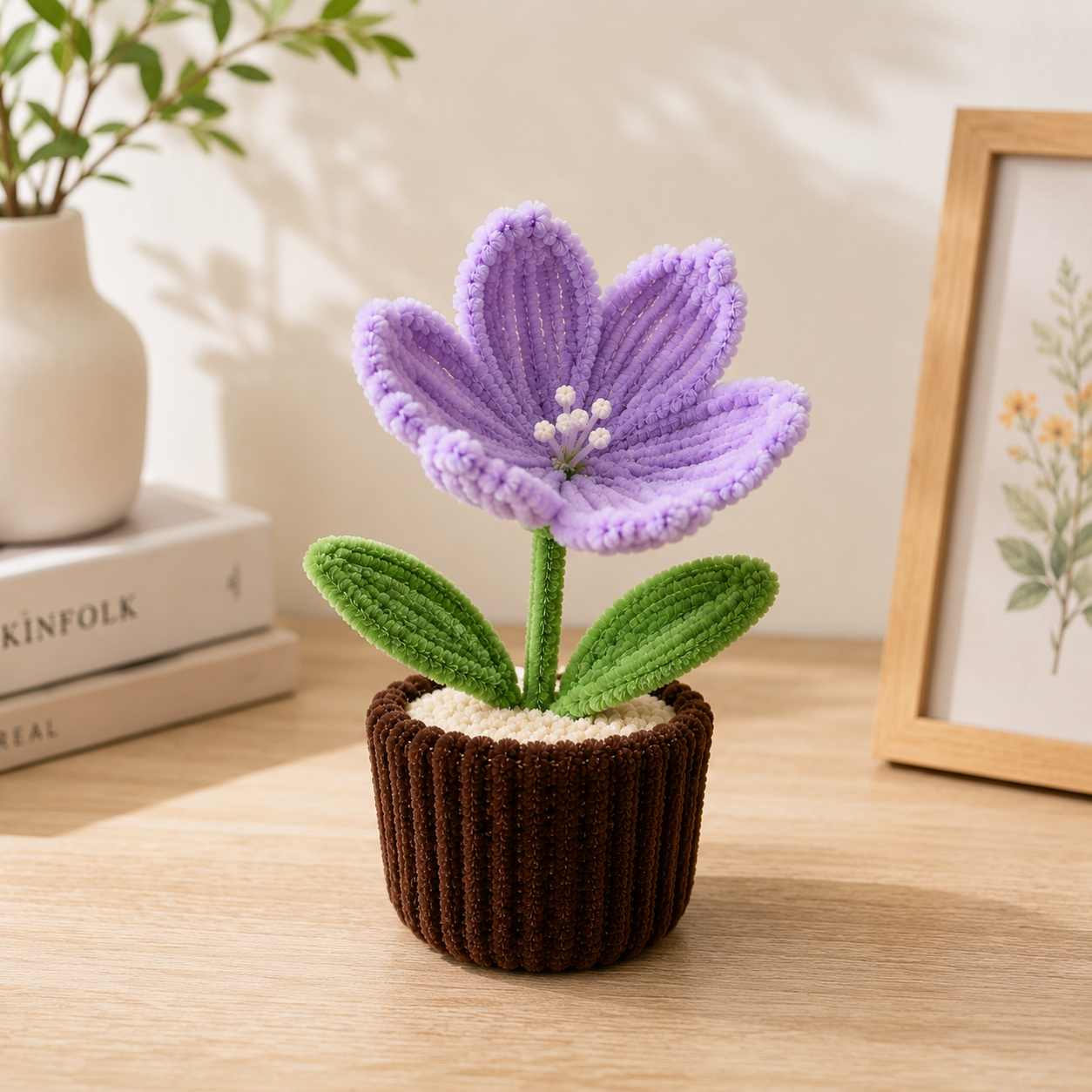 A crocheted purple flower with green leaves in a brown pot, displayed on a wooden table next to a framed botanical print and a small vase.