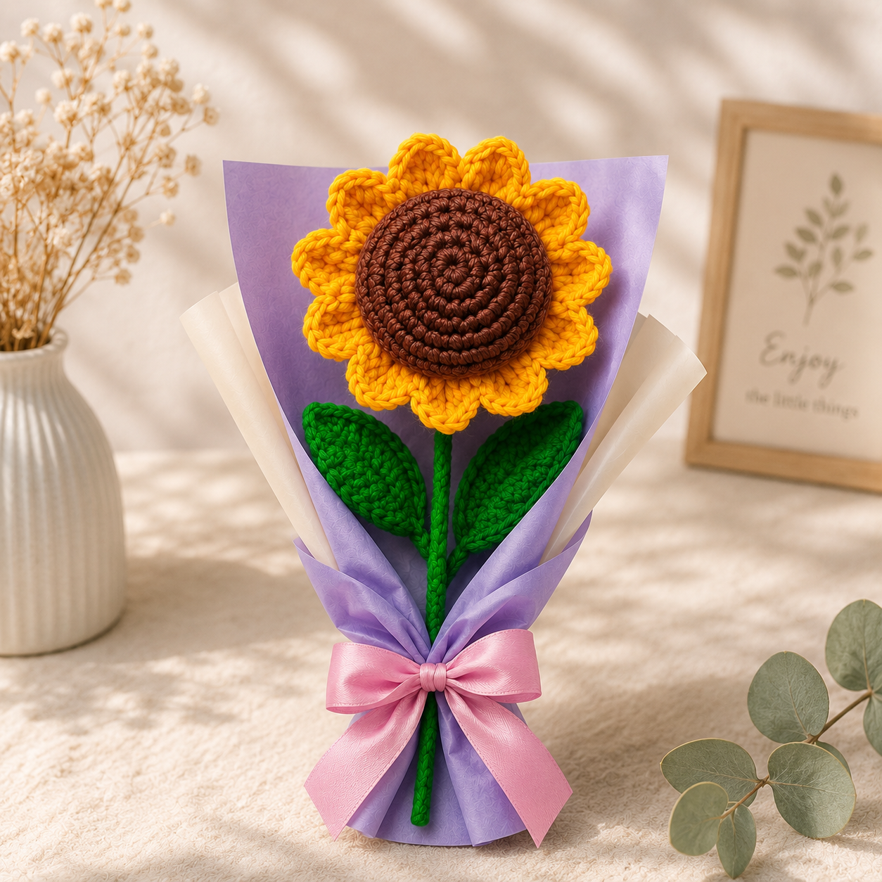 A crocheted sunflower gift bouquet with a brown and yellow crochet flower, green leaves, and a pink ribbon bow, wrapped in purple and white paper on a styled tabletop.