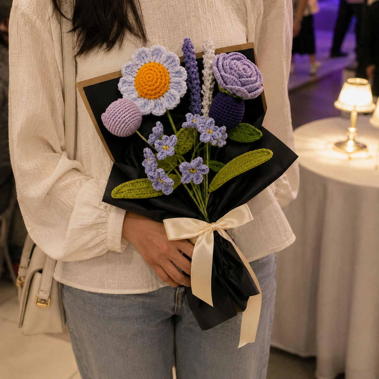 A person in a white ribbed sweater and light blue jeans holds a handmade crochet flower bouquet wrapped in black paper with a cream ribbon. The bouquet features purple, lavender, white, and yellow crocheted flowers with green leaves, set against a softly blurred indoor background with a lit table lamp.