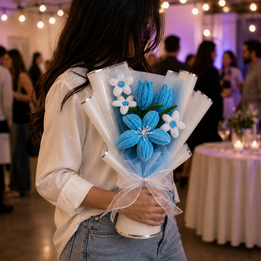 A person in a white shirt and jeans holds a handmade crochet flower bouquet with blue and white flowers wrapped in white tissue paper, set against a blurry event background with warm string lights and tables.