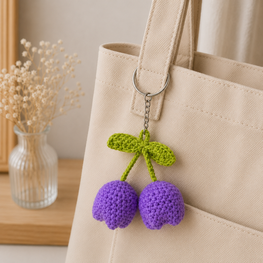 A beige canvas tote bag with a purple and green crocheted cherry keychain hanging from the handle, shown next to a small glass vase with dried flowers on a wooden surface.