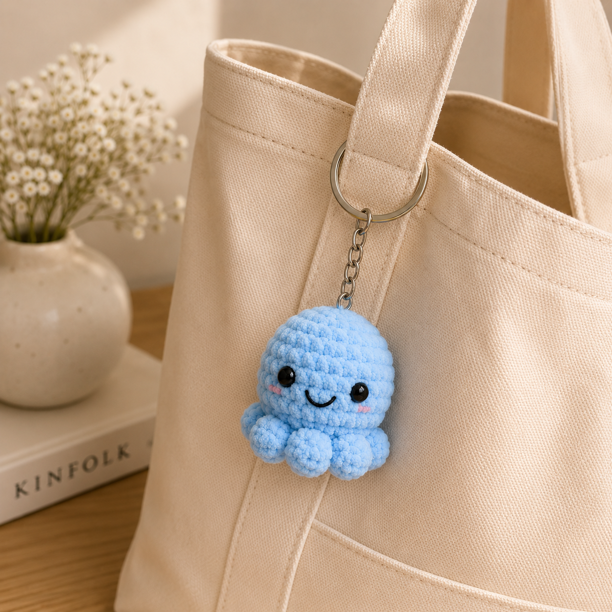 A light blue crocheted octopus keychain charm with a smiling face hangs from a silver metal ring attached to a beige canvas tote bag. The background features a warm, softly lit setting with a small vase of baby's breath and a book.