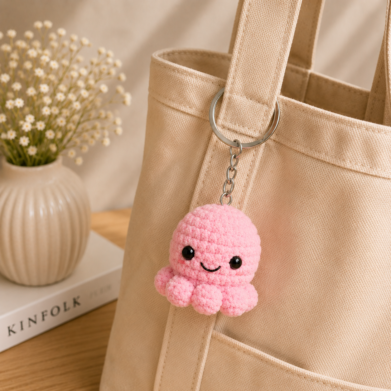 A pink crocheted octopus keychain charm hanging from a silver metal ring on a beige canvas tote bag, styled next to a small vase of baby's breath on a wooden surface.