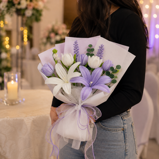 A person in a black long-sleeve top and light blue jeans holds a wrapped bouquet of white and lavender lilies with green foliage in a softly lit indoor setting with blurred lights and tables in the background.