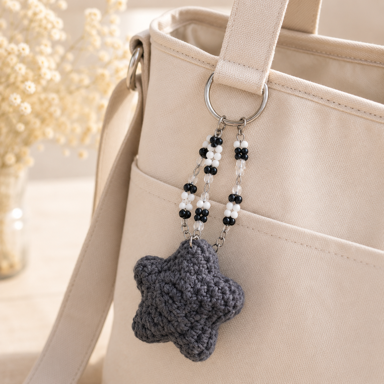 A close-up of a beige canvas tote bag with a dark gray crocheted star charm keychain hanging from a silver ring. The keychain features black and white beaded strands.