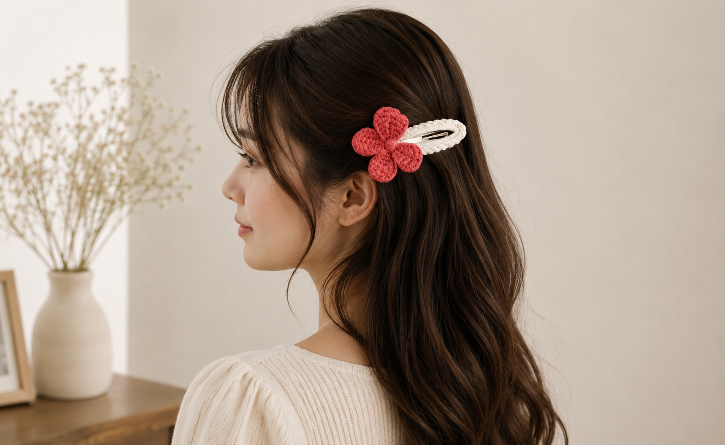 A woman with long dark brown hair is shown in profile wearing a pink crocheted bow hair clip with a white elastic band, set against a soft beige background with a vase of dried flowers on a table.