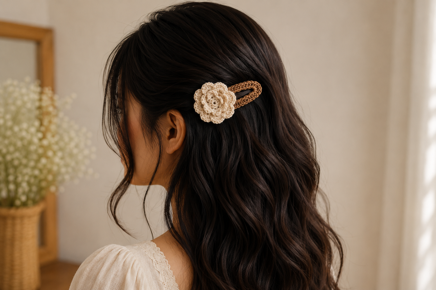 A woman with long, wavy dark brown hair is shown from the side, wearing a cream-colored crochet flower hair clip with a gold-toned metal band. The background is a softly lit, warm indoor setting with a blurred mirror and dried flowers.