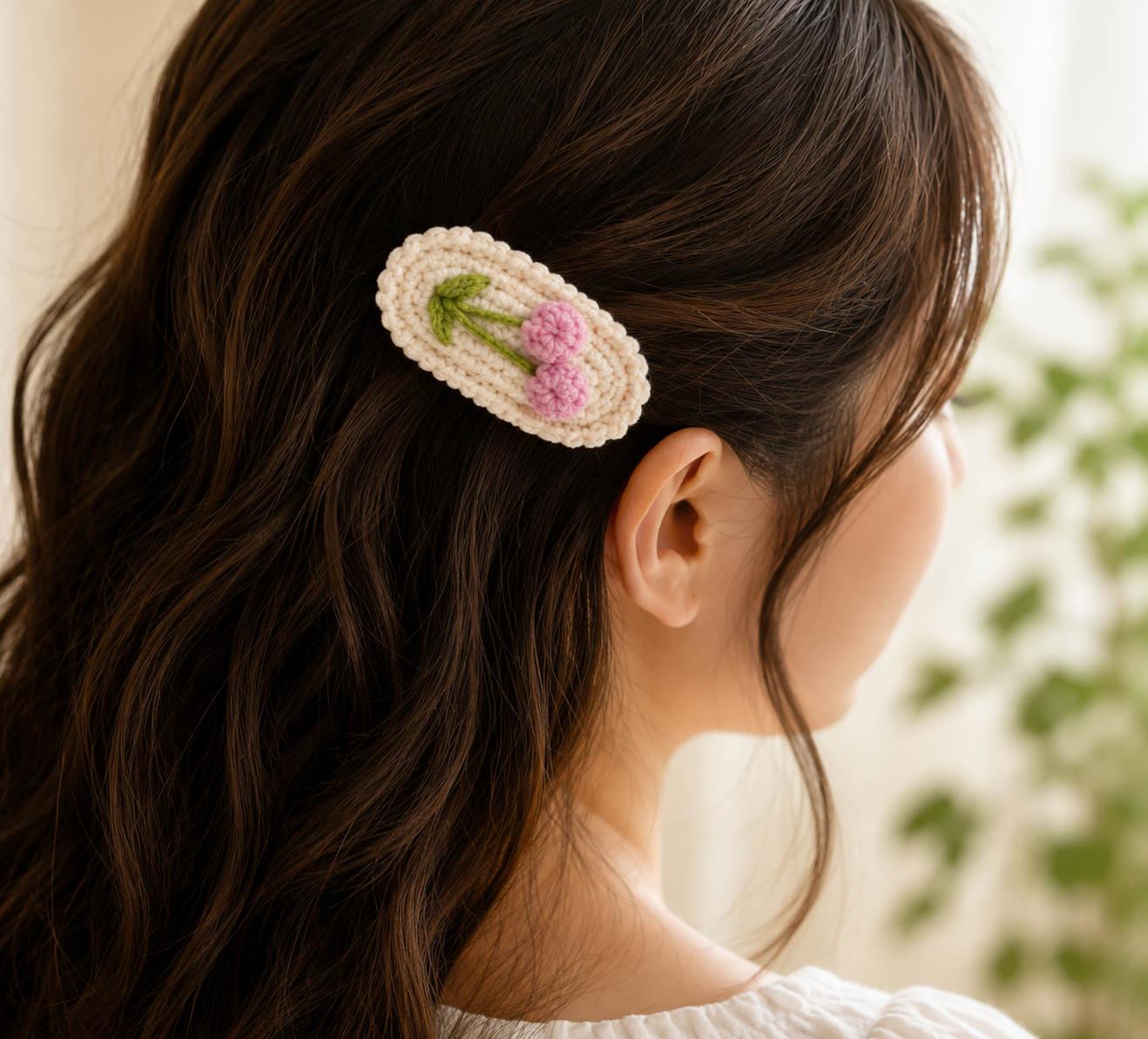 A close-up side view of a woman with long dark brown hair wearing a crocheted hair clip. The clip is cream-colored with a pink flower and green leaf design, set against a softly blurred indoor background.