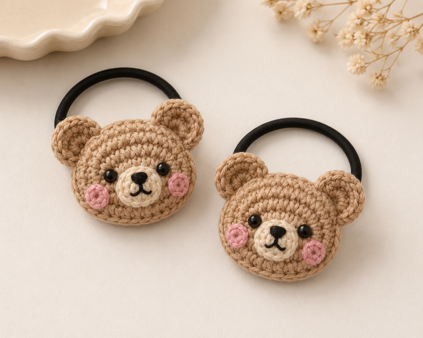 Two black elastic hair ties shaped like crocheted teddy bear heads, displayed on a light beige surface with dried flowers and a ceramic dish in the background.