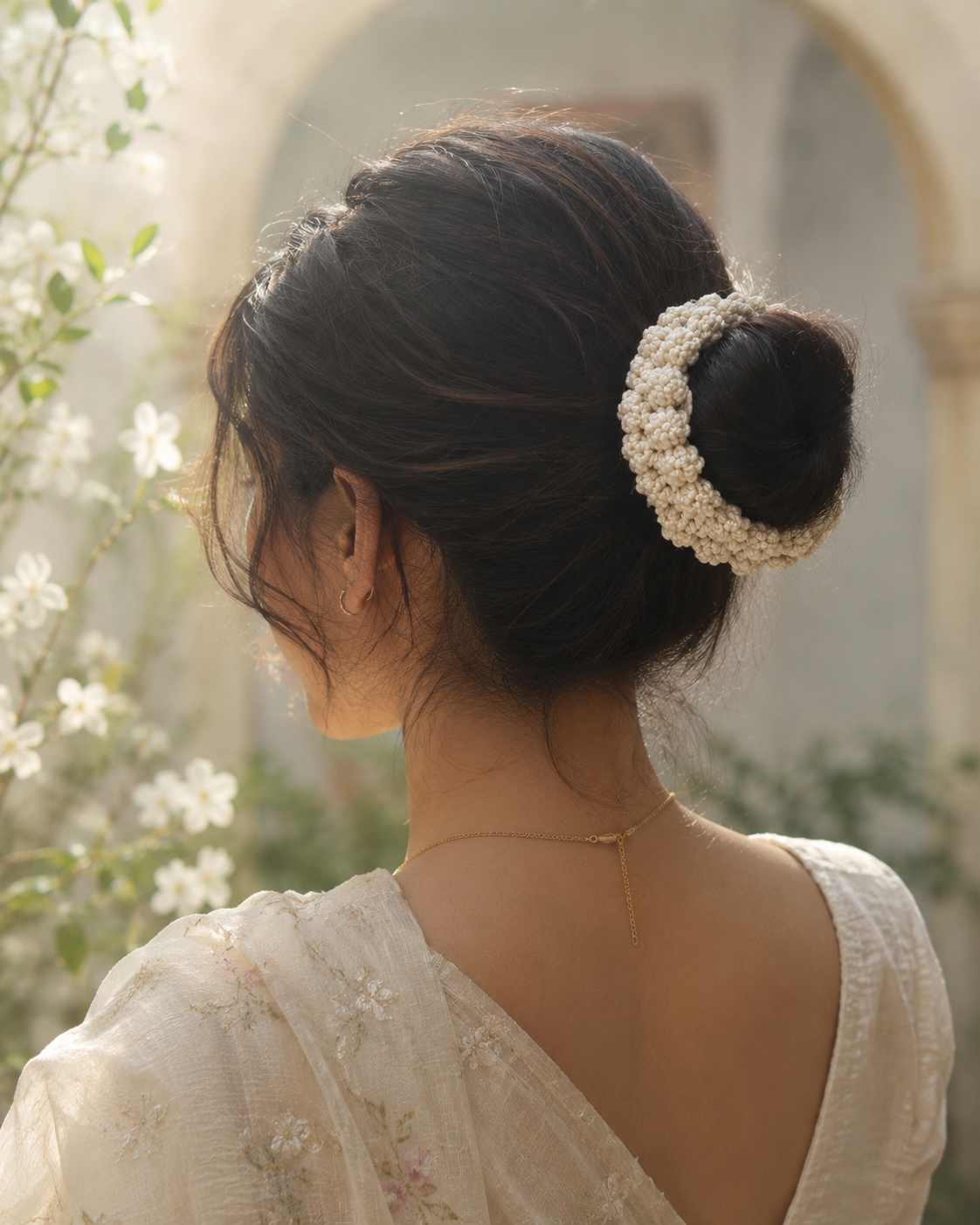 A woman shown from the back wearing a cream-colored, pearl-like beaded hair bun accessory in her dark hair, set against a soft-focus outdoor background with white flowers and a stone archway.