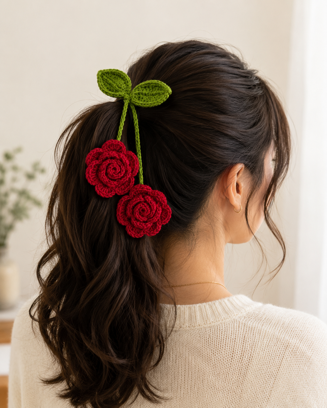 A woman shown from the back wearing a dark brown ponytail with a handmade crochet hair accessory featuring two red roses and green leaves, styled with a cream knit sweater in a bright indoor setting.