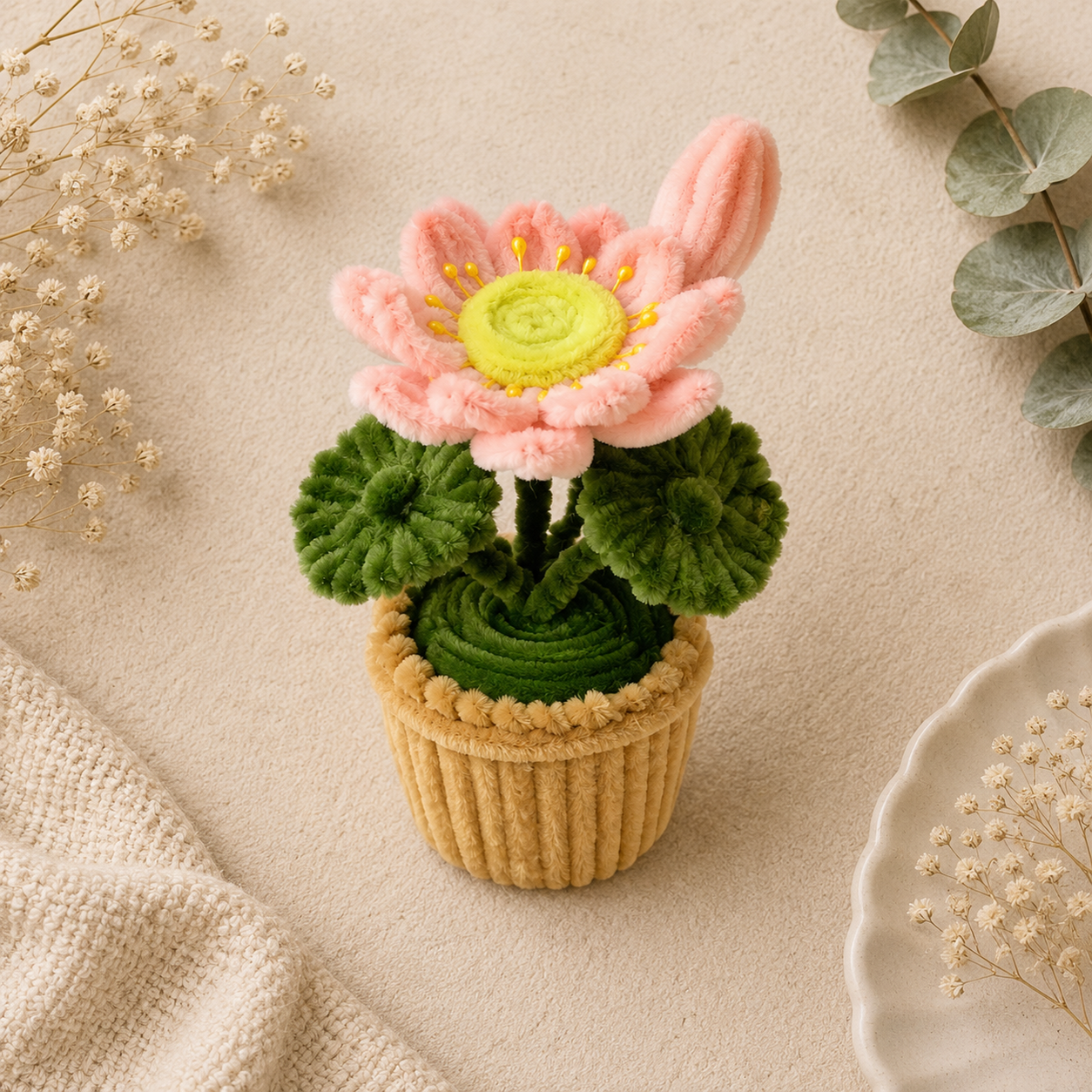 A handmade plush flower pot with a pink flower, green leaves, and a beige pot, styled on a neutral beige background with dried flowers and eucalyptus.